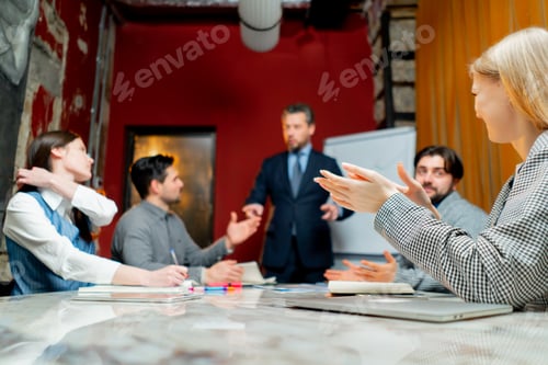 Preview: Office workers sit in a conference room with red walls and director explains the drawn up schedule