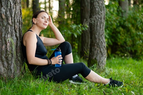 Preview: Beautiful young woman resting by tree after forest run, holding a water bottle and enjoying nature