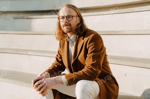 Preview: Ginger man wearing eyeglasses drinking coffee while sitting on stairs