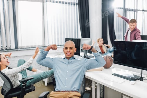 Preview: happy young african american man showing biceps and smiling at camera while colleagues having fun