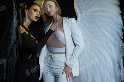 Preview: women in costumes of dark demon and light angel with fantasy wings looking at camera on black