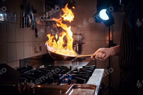 Preview: Cooker wearing gloves and apron frying flambe on a pan in a dark restaurant kitchen