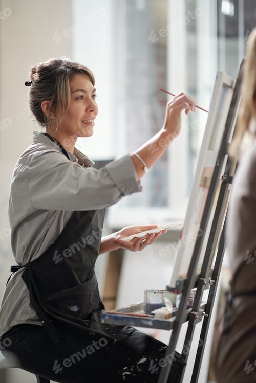 Preview: Happy young smiling female with paintbrush and palette working over picture