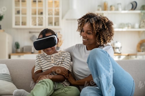 Preview: Happy joyful African American family mother and son using VR headset while relaxing together on sofa