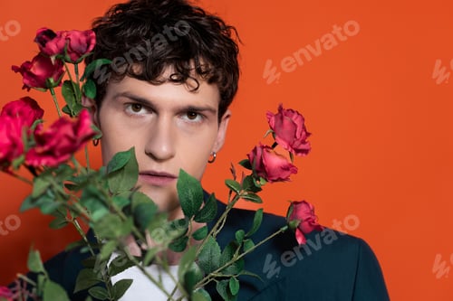 Preview: Portrait of curly man in jacket looking at camera near flowers for 8 march holiday on red background