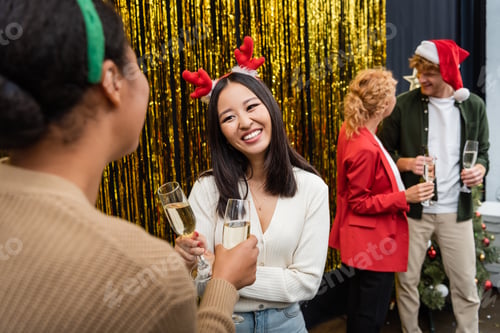 Preview: Smiling asian businesswoman holding champagne and talking to multiracial colleague during Christmas