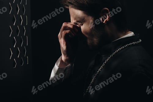 Preview: young tense catholic priest touching face with closed eyes near confessional grille in dark with