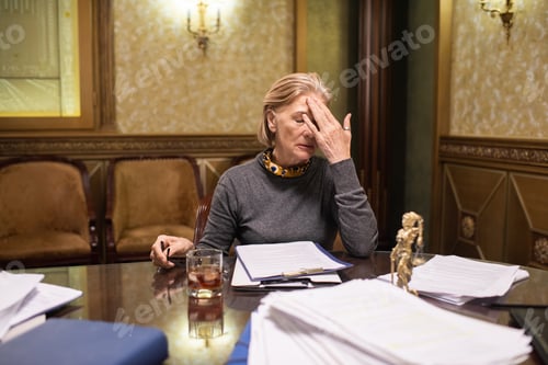 Preview: Blond mature female lawyer touching her head while working overtime