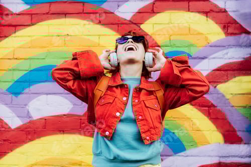Preview: Emotional stylish woman wearing wireless headphones, listening to music and singing on rainbow wall