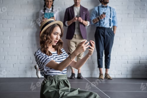 Preview: young stylish woman taking selfie on smartphone while her friends using digital devices