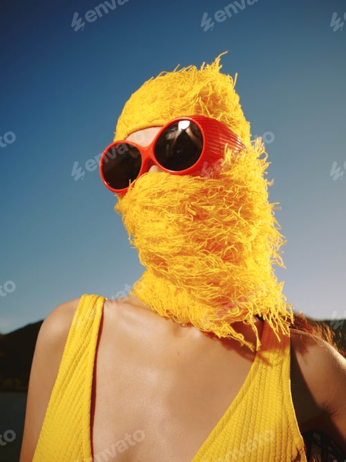 Preview: Woman in yellow dress with sunglasses and hat featuring a feather, posing outdoors on a sunny day