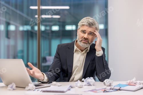 Preview: Stressed businessman sitting at desk in office with papers and laptop