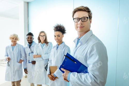 Preview: Multiracial group of smiling medical interns in lab coats standing in a row with clipboards and
