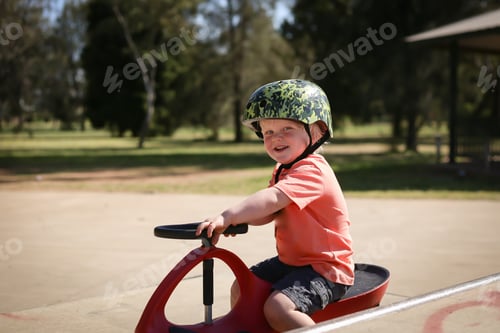Preview: Adorable blonde Australian kid with a helmet happily riding a children's car in the playground
