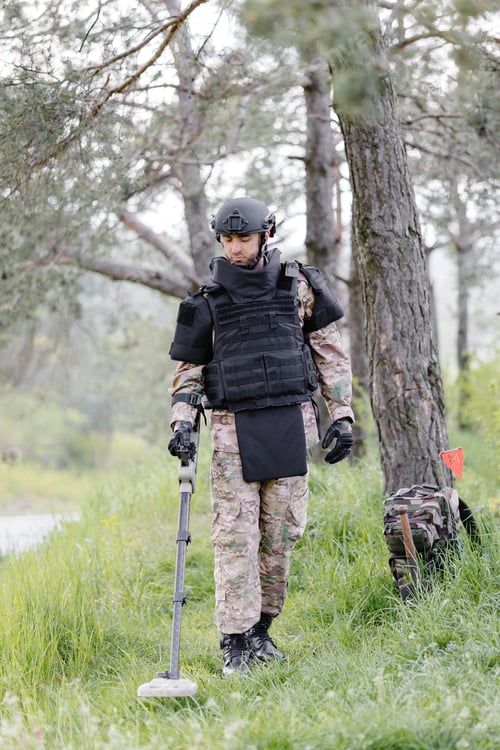 Preview: A man in a military uniform and bulletproof vest works in the forest with a metal detector