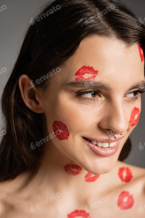 Preview: portrait of happy woman with piercing and red lipstick marks on face looking away isolated on grey
