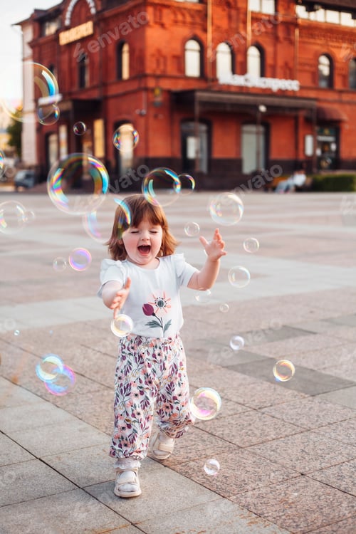 Preview: little girl playing fun with soap bubbles on the street in the city in summer at sunset
