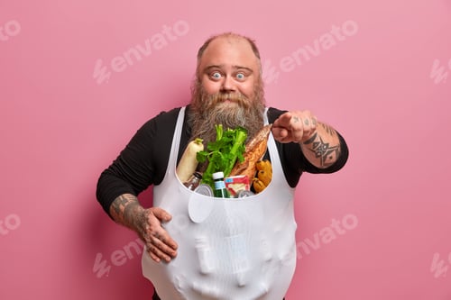 Preview: Surprised chunky bearded thick man indicates index finger at camera, has overweight as eats junk foo
