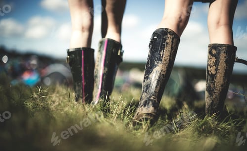 Preview: Low section rear view of two young women at a summer music festival wearing muddy Wellington boots.