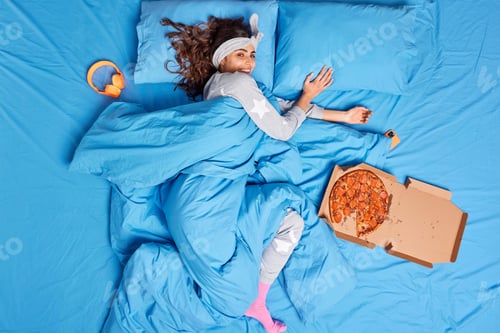 Preview: Top view of happy young female model lying in bed under warm blue blanket spends lazy day at home ea