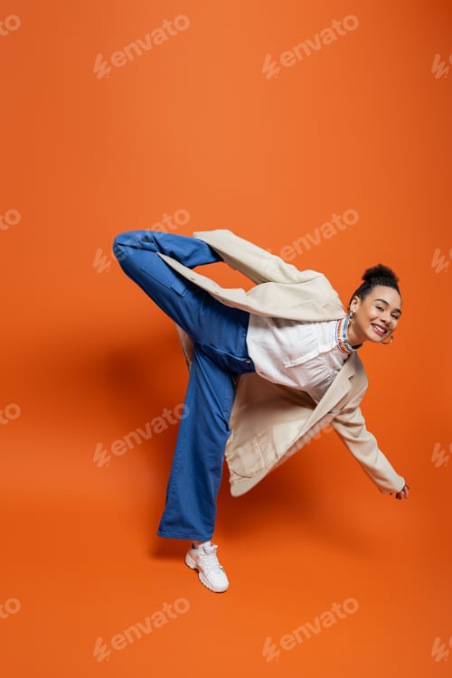 Preview: jolly african american model in stylish urban outfit holding playfully raised leg behind her back