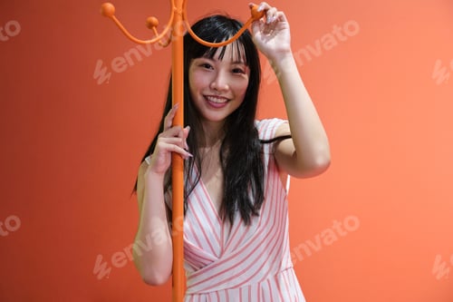 Preview: Young asian woman pose with a red orange monochrome coat rack. Studio shot.