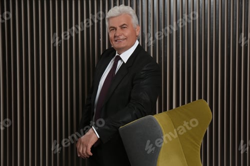 Preview: Happy mature businessman standing near armchair near wood slat wall