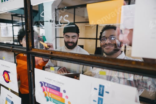 Preview: Smiling colleagues working on plan of project in workspace with stickers on glass wall