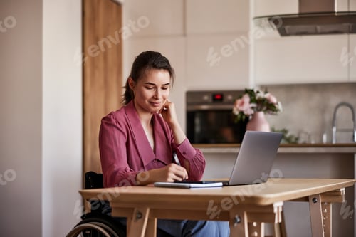 Preview: Portrait of a smiling woman in wheelchair taking notes at wooden kitchen table with laptop.