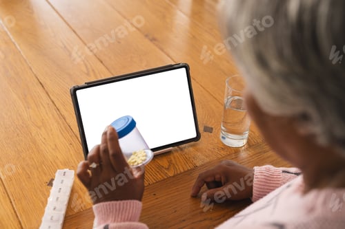Preview: Senior biracial woman having video call with doctor on tablet with copy space, holding pills at home