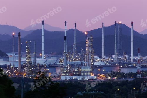 Preview: An illuminated oil refinery complex set against mountain silhouettes at dusk
