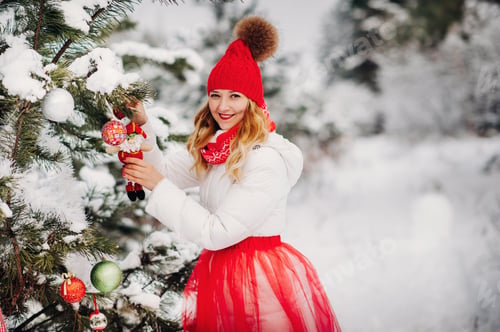 Preview: A girl in a white jacket and red hat dresses up a Christmas tree in the forest in winter.A girl