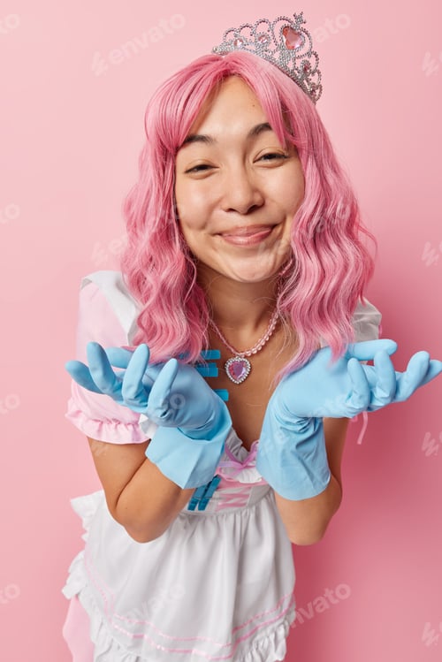 Preview: Happy Asian woman with long dyed pink hair spreads palms wears dress crown and rubber gloves does do
