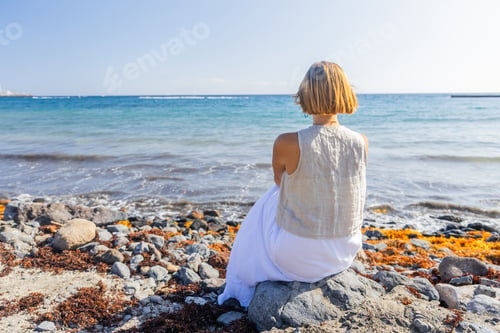 Preview: A woman sits on a rock by the ocean, looking out at the water