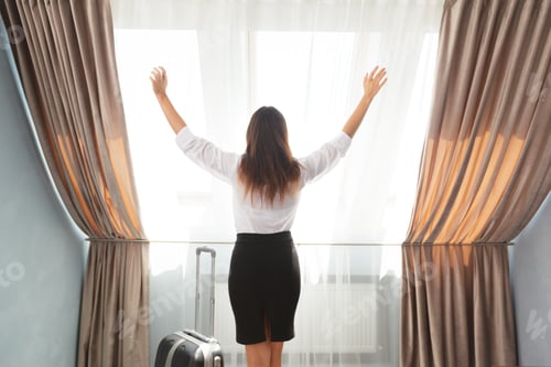 Preview: Woman stretches her arms in a hotel room while welcoming the morning light through the curtains