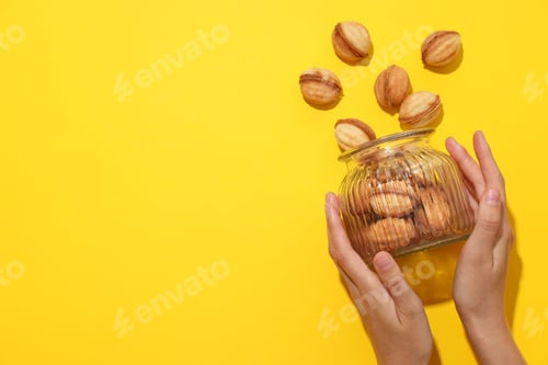 Preview: Cookie nuts in glass jar in female hands on yellow background, space for text