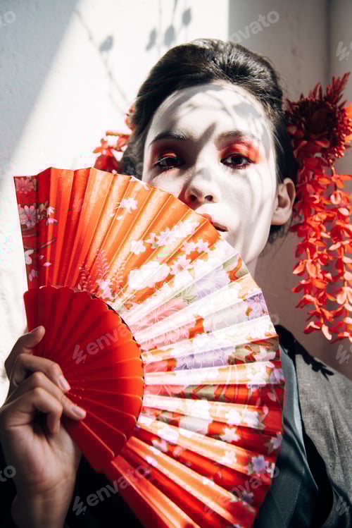 Preview: portrait of beautiful geisha with red and white makeup holding hand fan in sunlight