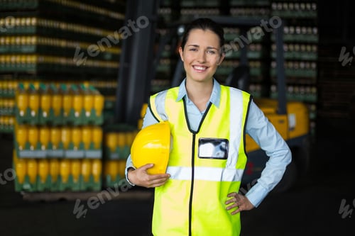 Preview: Female warehouse supervisor standing in storage wearing safety vest holding hardhat with forklift