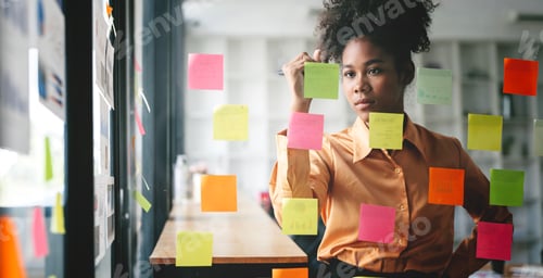 Preview: Young businesswoman using sticky notes in glass wall to writing strategy business plan to success