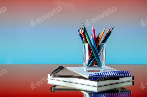 Preview: Side view of colored pencils kept in a glass jar on stacked spiral notebooks on the left side on