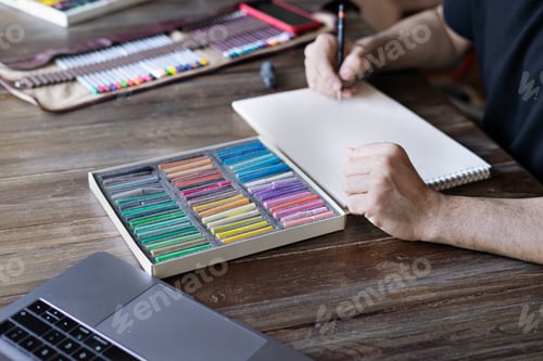 Preview: man artist painting with pencil and pastel crayon chalks on paper in front of laptop