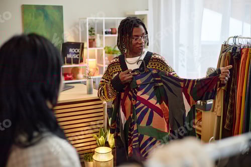 Preview: Young Adult Black Man Showing Second Hand Clothing to Customer Indoors