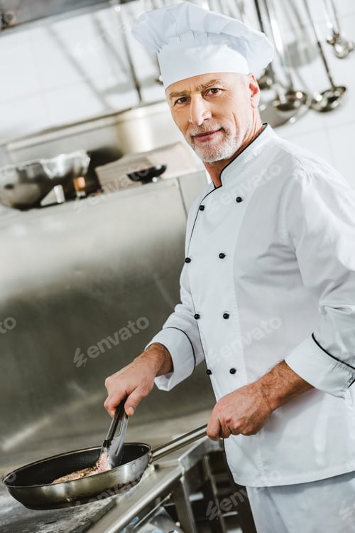 Preview: male chef looking at camera while roasting meat steak in restaurant kitchen