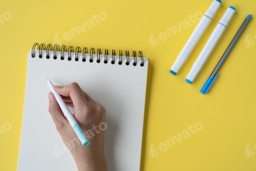 Preview: High angle shot of a hand writing in a journal with a marker pen on a yellow background