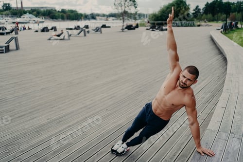 Preview: Self determined muscular man dressed in sport clothes stands in side plank, keeps arm raised