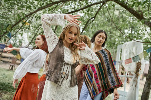 Preview: trendy young blonde woman dancing and looking at camera near friends in retreat center