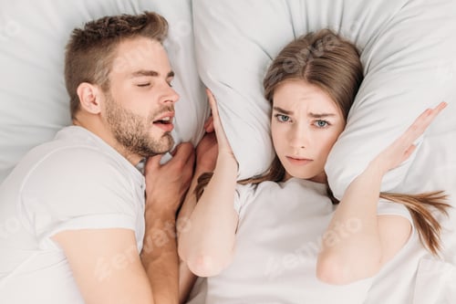 Preview: top view of woman plugging ears with pillow while lying in bed near snoring husband