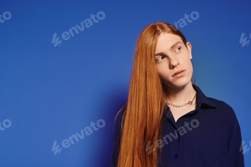 Preview: Young queer man with long red hair showcases stylish fashion against a vibrant blue backdrop