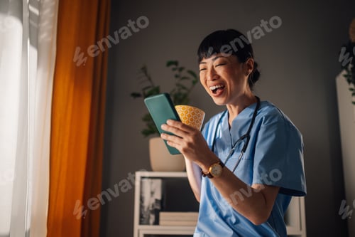 Preview: Asian woman nurse standing by window, with phone and yellow patterned mug