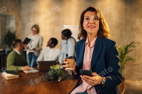 Preview: Smiling mature businesswoman with coffee and phone in hands on a meeting